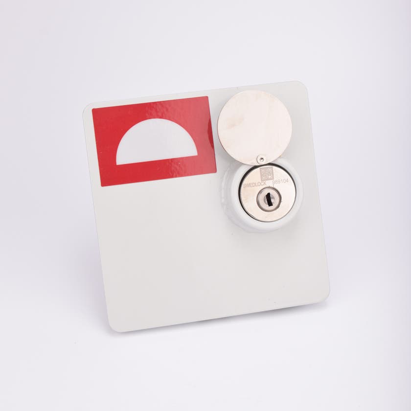 En vit fyrkantig brandlucka, med en rektangulär röd dekal i vänstra hörnet. Luckan har ett metalliskt lås med nyckelhål.