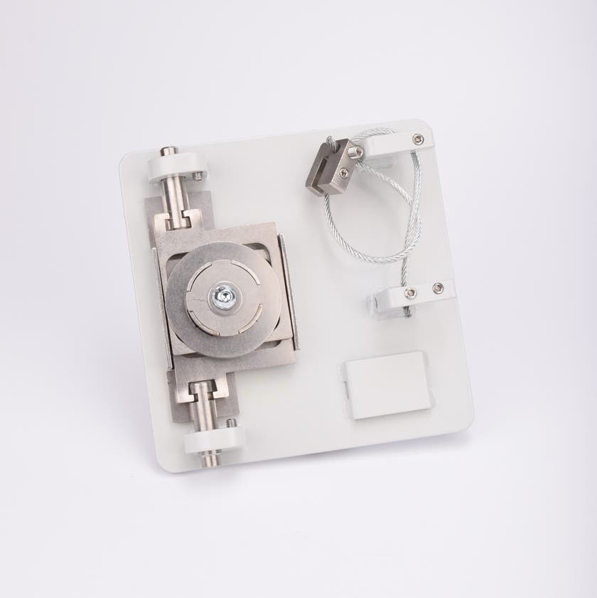 Metallkomponent monterad på vit bas med centralt bultfäste, två cylindrar, trådögla och klämmor.