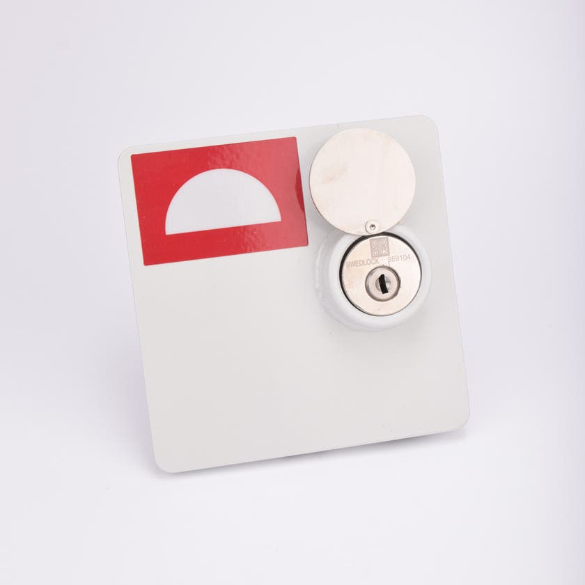 En vit fyrkantig brandlucka, med en rektangulär röd dekal i vänstra hörnet. Luckan har ett metalliskt lås med nyckelhål.