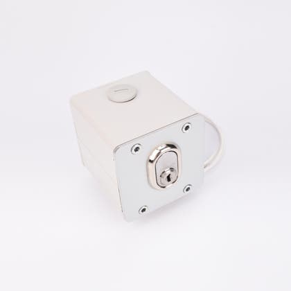 Vit fyrkantig elektrisk nyckelbrytare med metallisk låscylinder, rund knapp upptill, fyra hörnskruvar och kabelutgång på sidan.