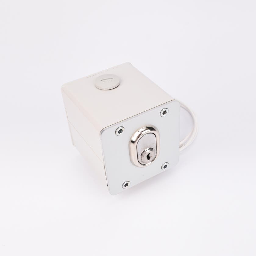 Vit fyrkantig elektrisk nyckelbrytare med metallisk låscylinder, rund knapp upptill, fyra hörnskruvar och kabelutgång på sidan.