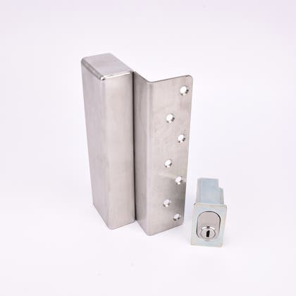 Två metallföremål visas. Det större är en L-formad nyckelförvaringsenhet med flera cirkulära hål längs ena sidan. Det mindre är en rektangulär cylinderhållare med en oval cylinder.