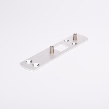 Rektangulär metallplatta med två cylindriska distanser och fyra försänkta hål, symmetriskt placerade. Plattan har även ett rektangulärt urtag i mitten.