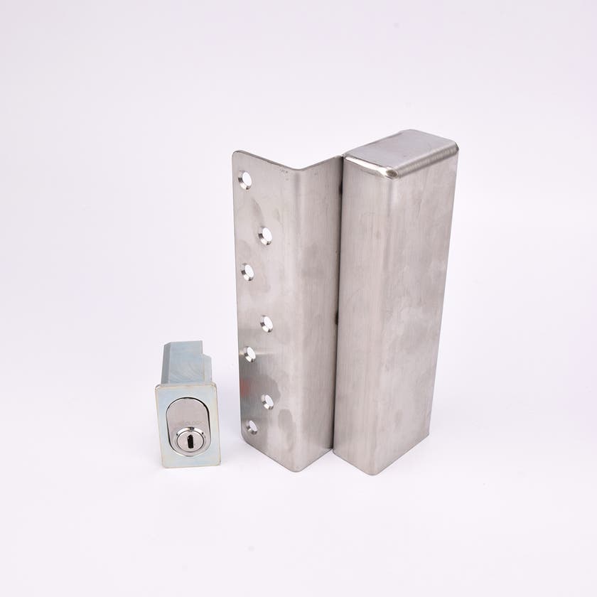 Två metallföremål visas. Det större är en L-formad nyckelförvaringsenhet med flera cirkulära hål längs ena sidan. Det mindre är en rektangulär cylinderhållare med en oval cylinder.