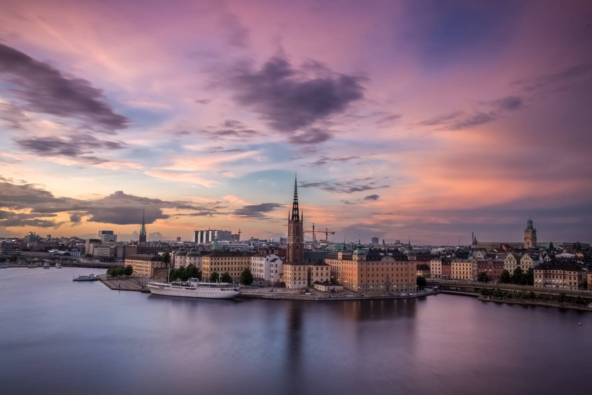 En bild över en Stockholm från avstånd. Stadsbilden speglar sig i vattnet. Tonerna i bilden går i rosa och lila