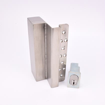 Två metallföremål visas. Det större är en L-formad nyckelförvaringsenhet med flera cirkulära hål längs ena sidan. Det mindre är en rektangulär cylinderhållare med en oval cylinder.