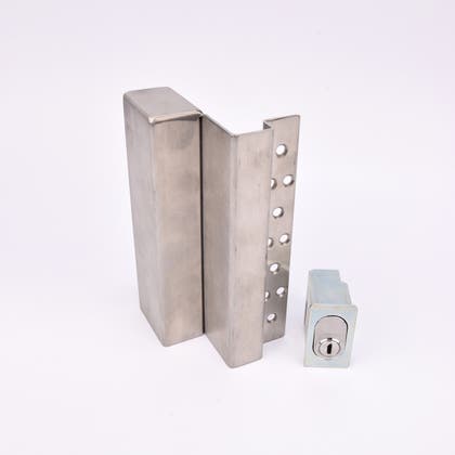 Två metallföremål visas. Det större är en L-formad nyckelförvaringsenhet med flera cirkulära hål längs ena sidan. Det mindre är en rektangulär cylinderhållare med en oval cylinder.