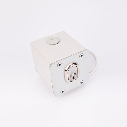 Vit fyrkantig elektrisk nyckelbrytare med metallisk låscylinder, rund knapp upptill, fyra hörnskruvar och kabelutgång på sidan.