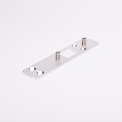 Rektangulär metallplatta med två cylindriska distanser och fyra försänkta hål, symmetriskt placerade. Plattan har även ett rektangulärt urtag i mitten.