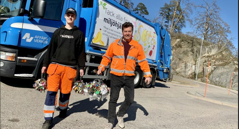 Två personer klädda i orange hållandes stora nyckelknippor framför en sopbil