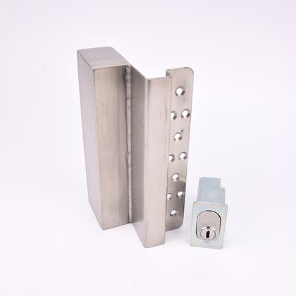 Två metallföremål visas. Det större är en L-formad nyckelförvaringsenhet med flera cirkulära hål längs ena sidan. Det mindre är en rektangulär cylinderhållare med en oval cylinder.